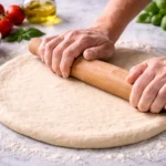 Rolling pizza dough on marble surface with wooden rolling pin and fresh ingredients in background