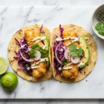 crispy fish tacos recipe with cod lime crema and slaw on white marble