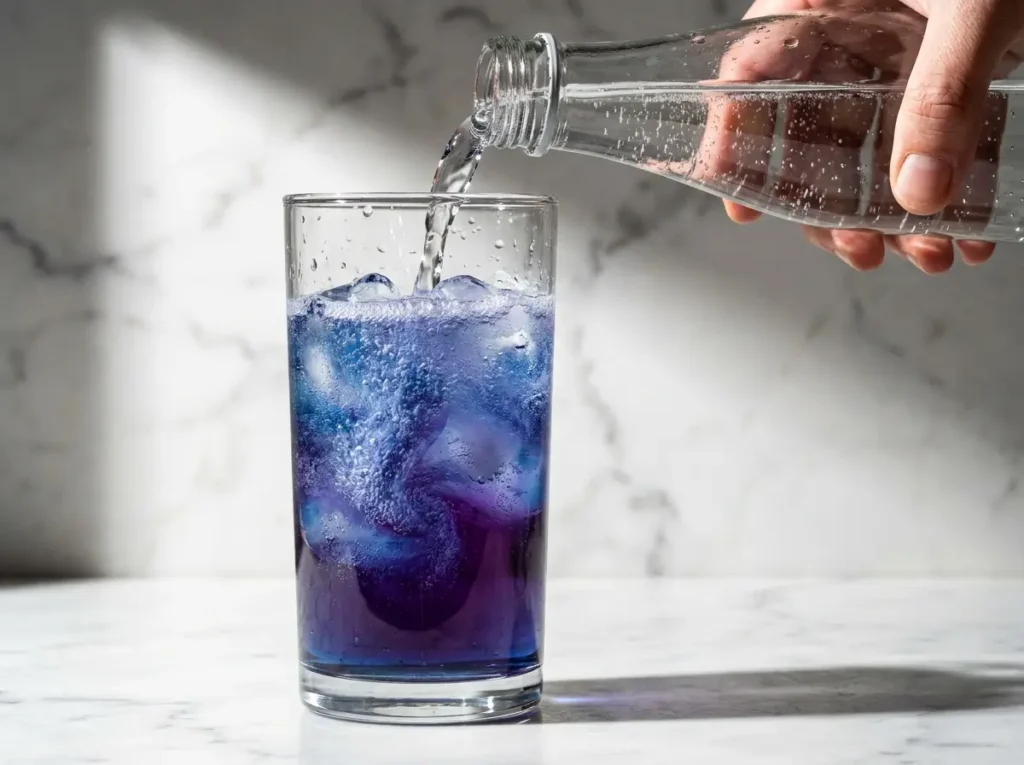  Hand pouring sparkling water into a blue lotus energy drink over ice, color shifting to purple