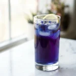A tall glass of homemade lotus energy drink with blue-purple gradient and lemon slice on white marble